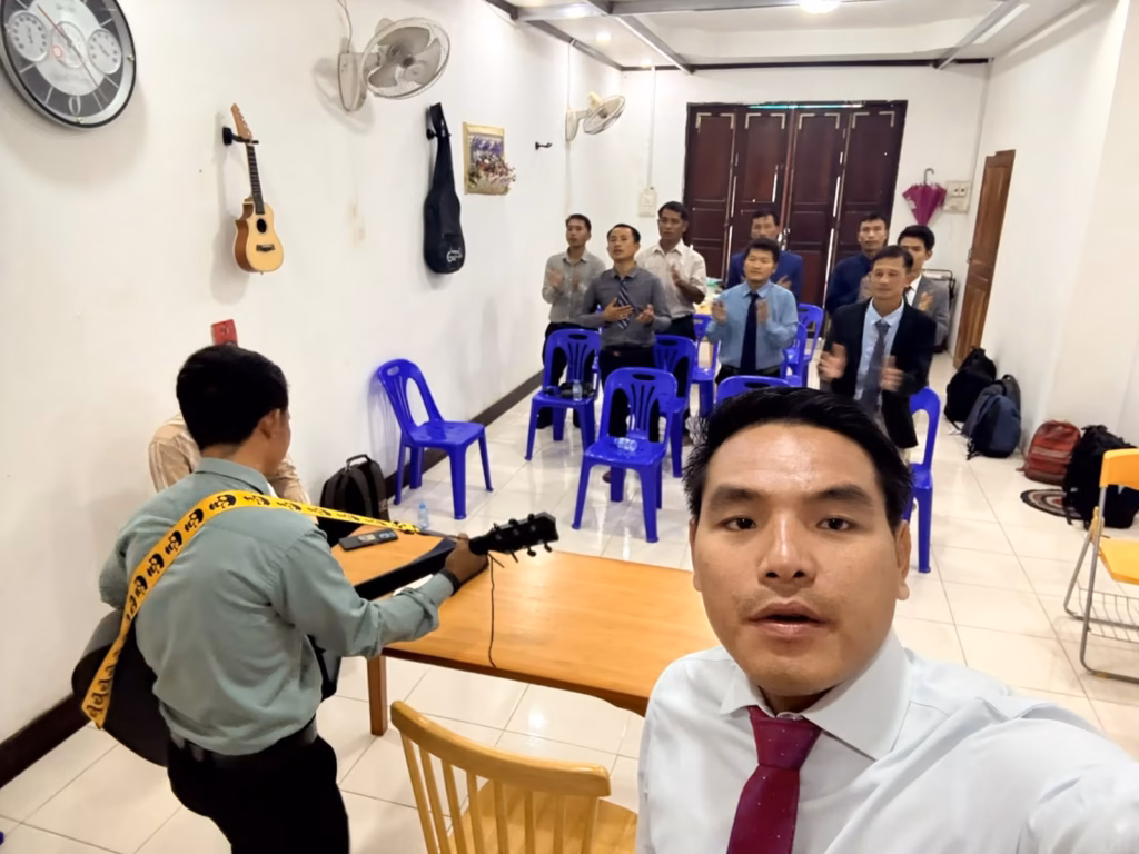 Pastor Khen taking a selfie in a tiled room while men behind him clap and sing and another man plays guitar in Laos.