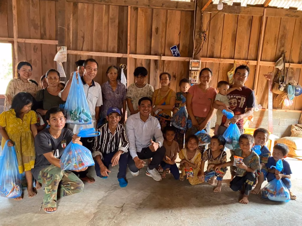 Pastor Khen in a white shirt crouching among villagers and children who hold blue bags of gifts inside a wooden house in Laos.