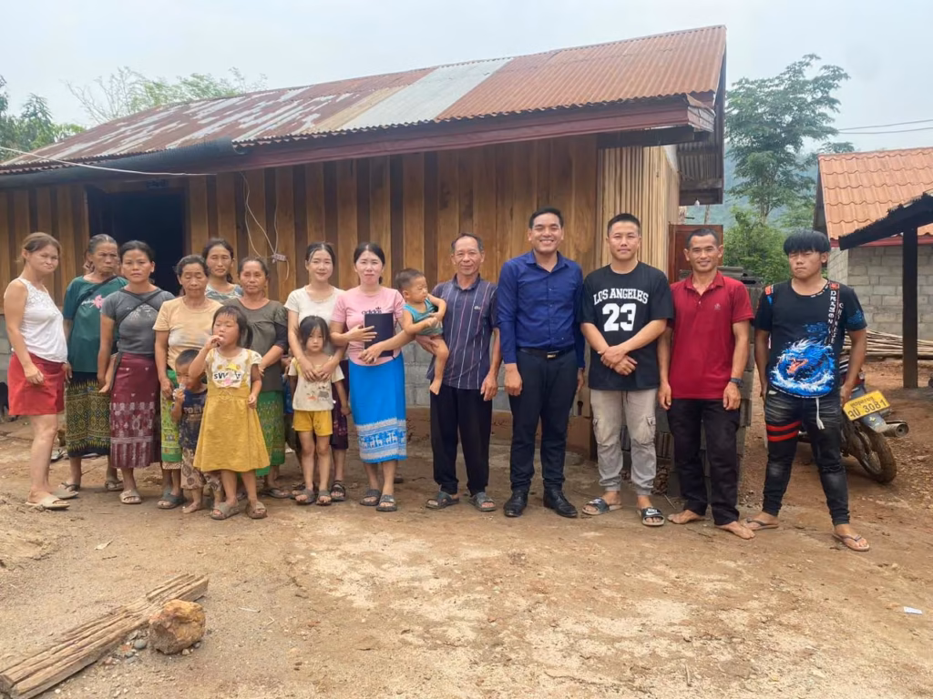 Pastor Khen in a blue shirt standing with villagers, including women and small children, in front of a wooden house with a tin roof in Laos.