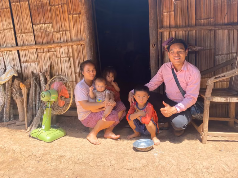 Pastor Khen in a pink shirt crouching with a thumbs up beside a mother and three young children at the doorway of a bamboo house in Laos.