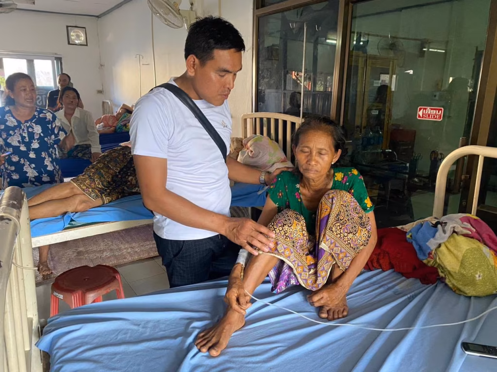 A pastor in a white shirt standing beside an older woman with an IV line sitting on a hospital bed, with other patients behind in Laos.