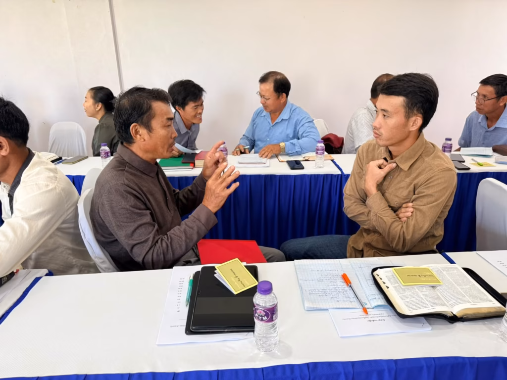 A pastor in a brown shirt gestures while talking with a man in a tan jacket over Bibles and notes at the pastors' conference in Vang Vieng, Laos.