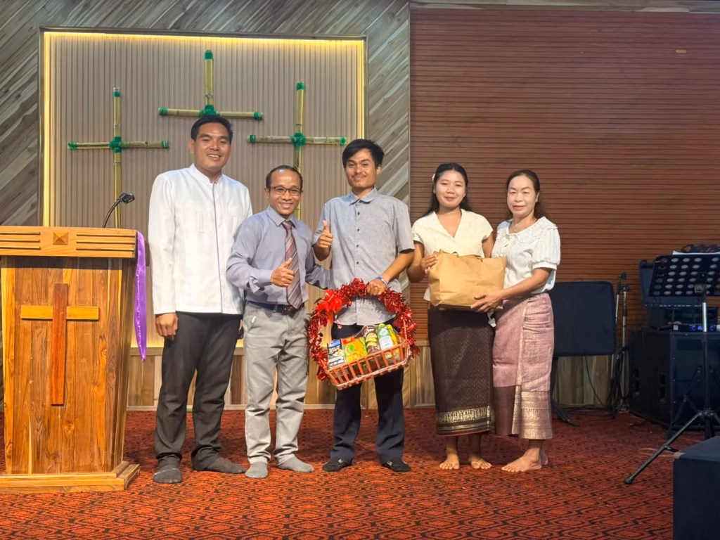 Pastor Khen and a fellow pastor posing with a man holding a red gift basket of snacks and two women holding a paper bag in front of bamboo crosses in a church in Laos.