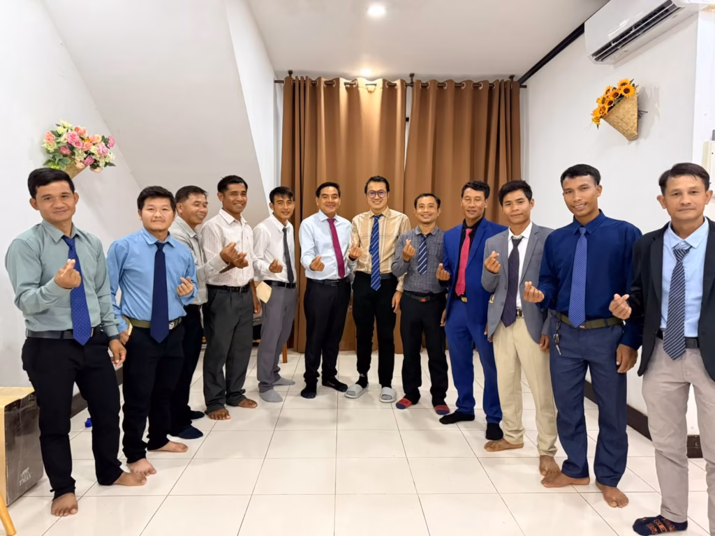 Pastor Khen posing with eleven other men in shirts and ties, each making a finger heart, in a tiled room with brown curtains in Laos.