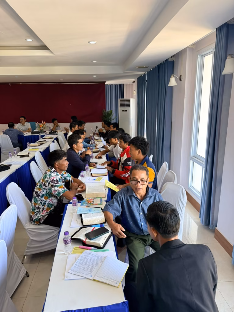A pastor in a blue shirt talks with a man in a black jacket along a long table where pairs of men discuss with Bibles at the pastors' conference in Vang Vieng, Laos.