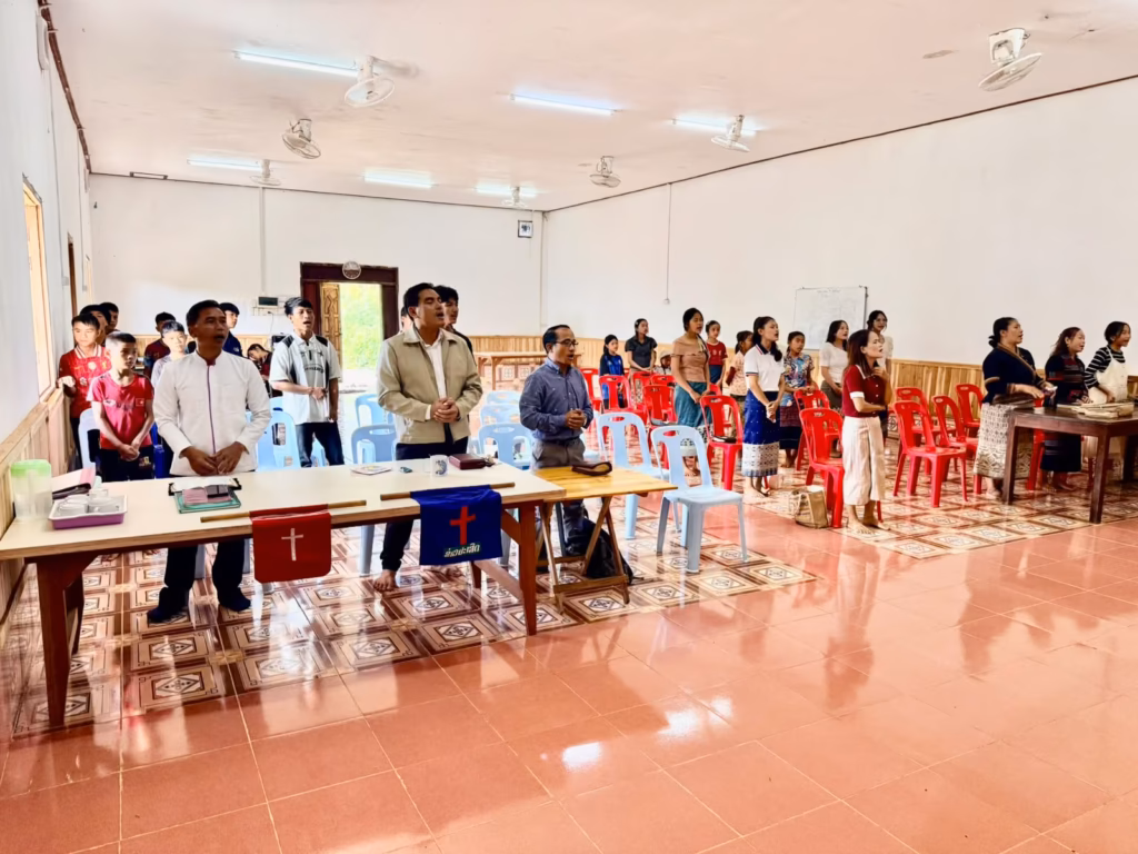 Pastor Khen and other men stand at a front table with red and blue cross banners while a congregation stands and sings in a large hall in Laos.