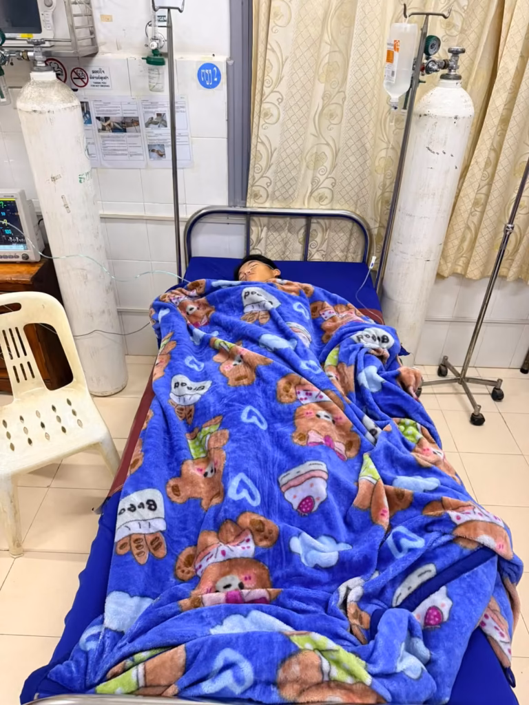 A boy from Pastor Khen's orphanage lying under a blue bear-print blanket in a hospital ward with oxygen tanks, an IV stand and a monitor beside the bed, after emergency surgery in Laos.