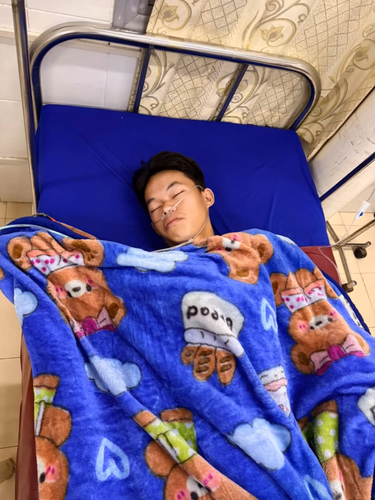 A boy from Pastor Khen's orphanage asleep in a hospital bed with an oxygen tube and an IV line, under a blue bear-print blanket, after emergency surgery for an appendix infection in Laos.