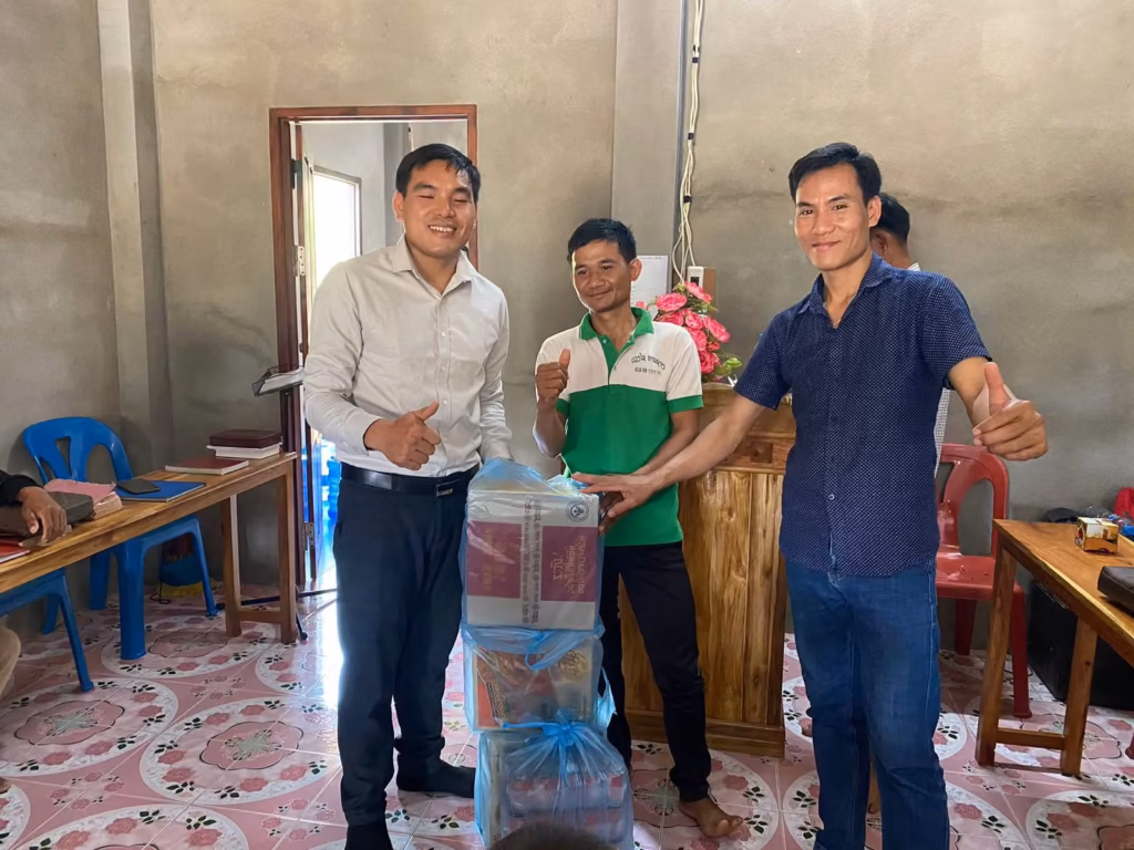 Pastor Khen standing with two other men giving thumbs up behind stacked boxes wrapped in blue plastic in a room with a wooden pulpit in Laos.