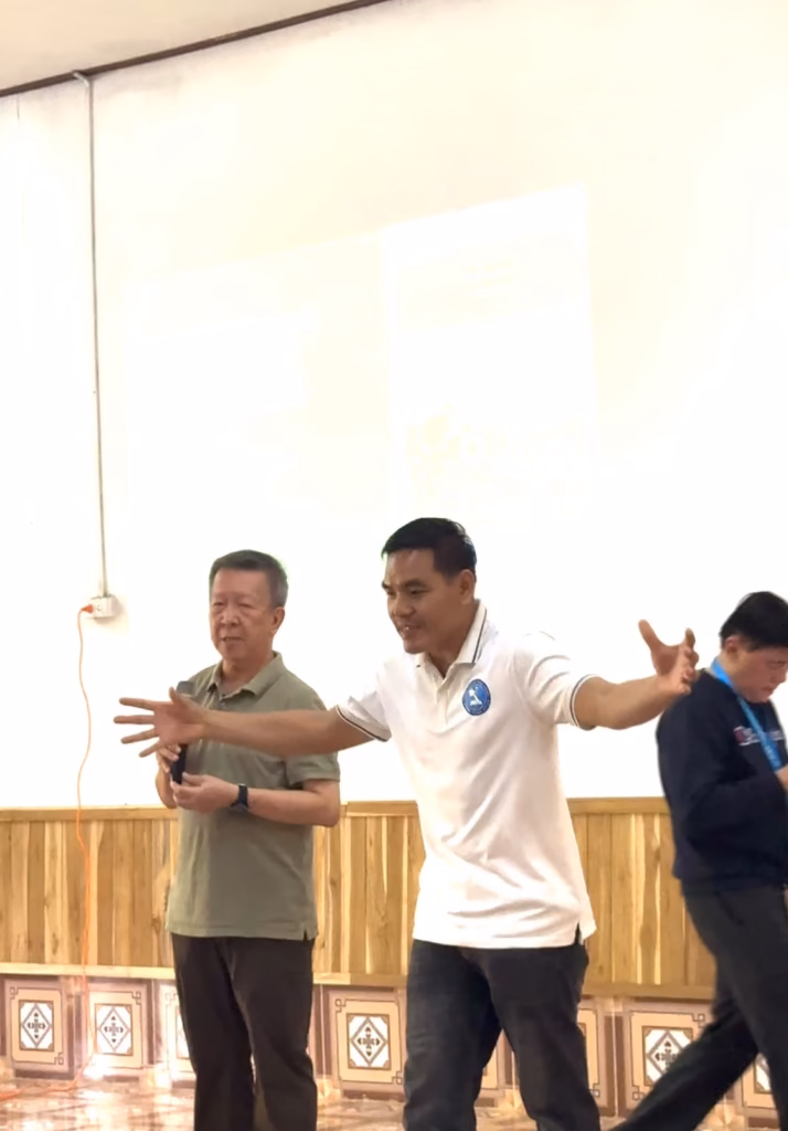 Pastor Khen, in a white polo shirt, standing with his arms outstretched in front of a projection screen while a fellow pastor in an olive shirt holds a microphone beside him in Laos.