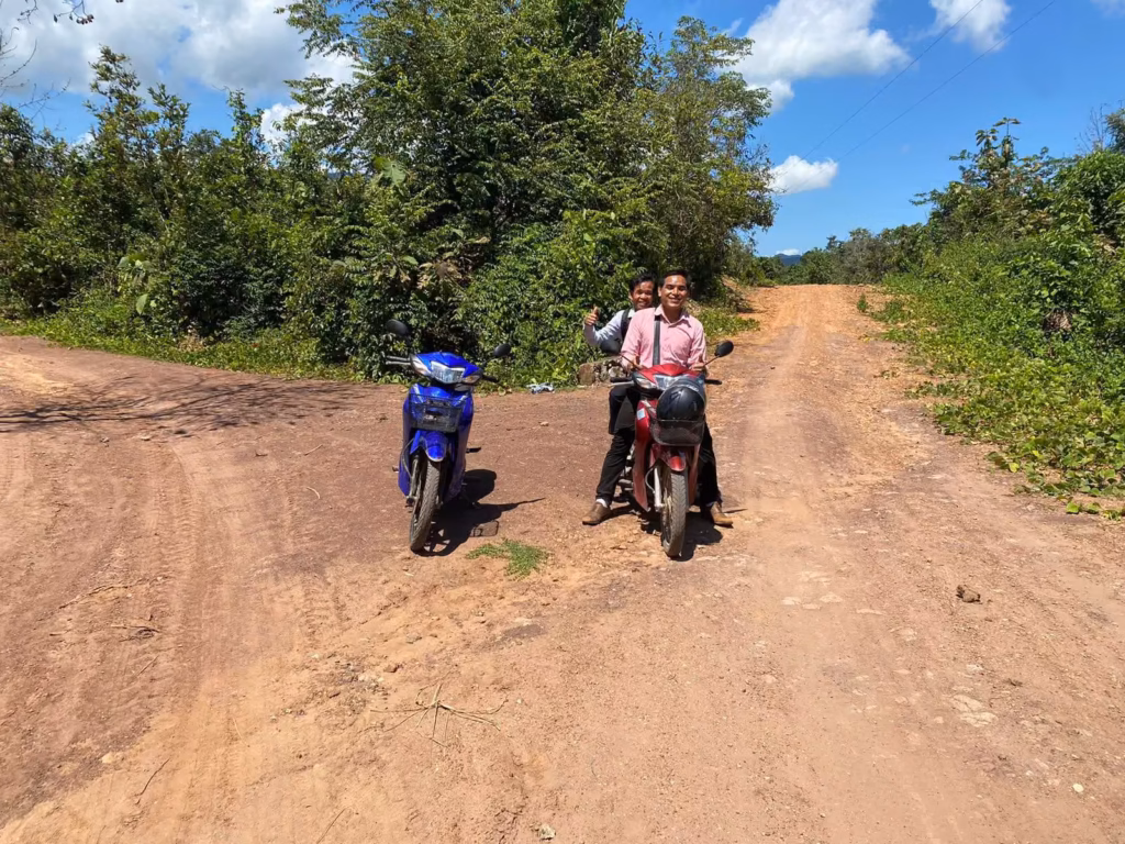 Pastor Khen and a fellow pastor riding together on a red motorbike on a dirt road beside a parked blue motorbike, with green hills behind in Laos.