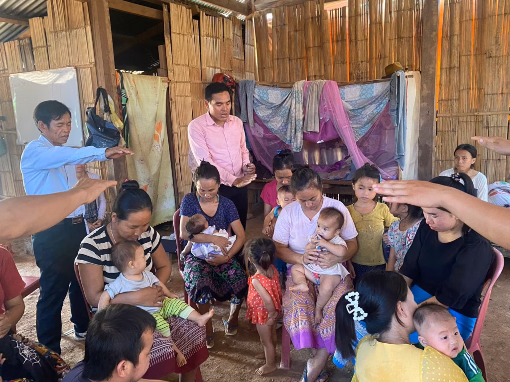 Pastor Khen and a fellow pastor standing with hands extended in prayer over seated mothers holding babies inside a bamboo house in Laos.