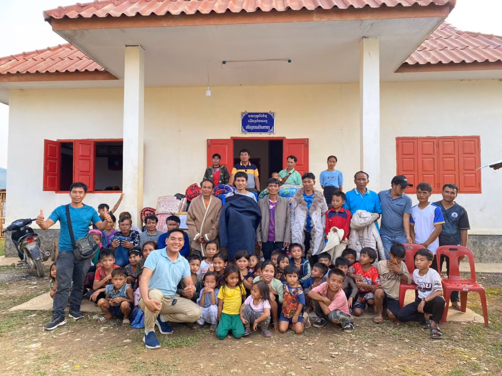 Pastor Khen crouching with a thumbs up in front of a large group of children and adults outside a white village office with red shutters in Laos.