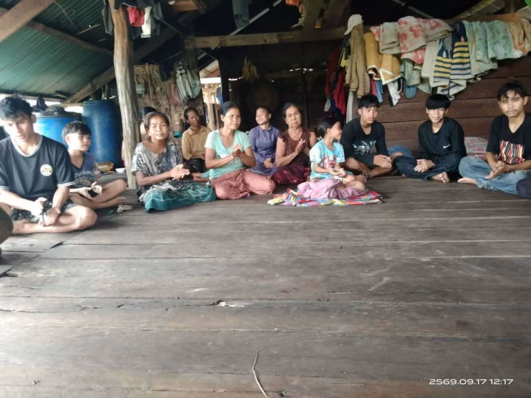 Women, teenagers and a young child sitting on a wooden floor and clapping inside a village house in Laos.