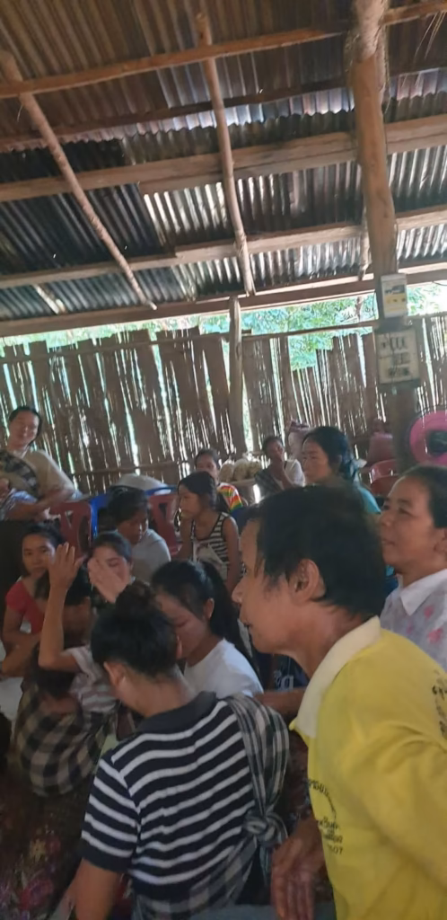 Adults and children seated closely together under a corrugated tin roof beside woven bamboo walls in Laos.