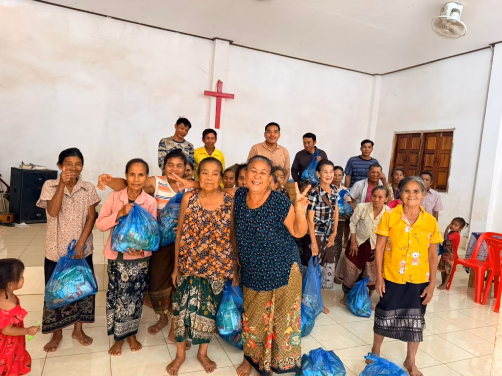 Women and children posing with blue gift bags on a tiled church floor while five men stand in the back row in Laos. Pastor Khen is pictured.