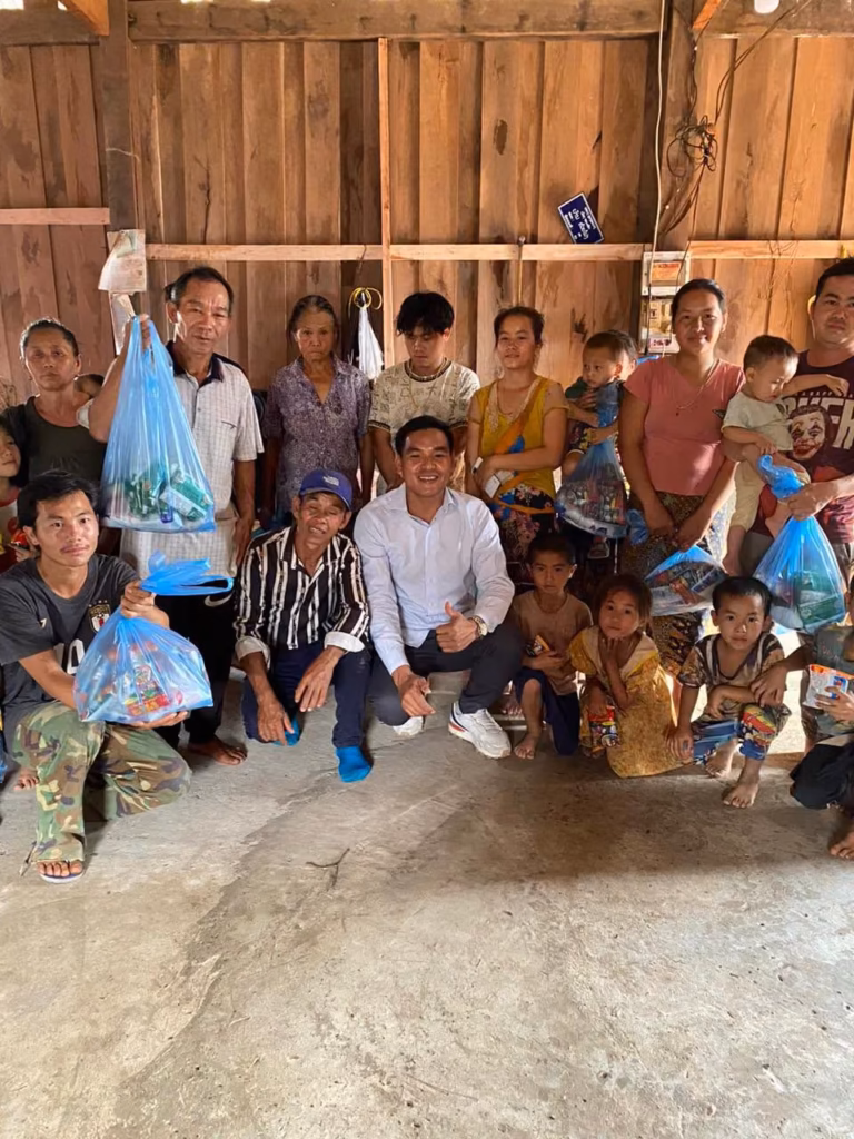 Pastor Khen in a white shirt crouching with a thumbs up among villagers holding blue gift bags on a concrete floor in Laos.