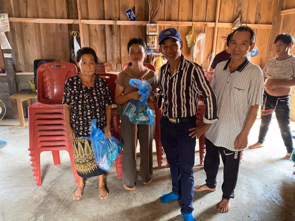 Two women and two men standing in front of stacked red chairs, two of them holding blue bags of goods, inside a wooden house in Laos.