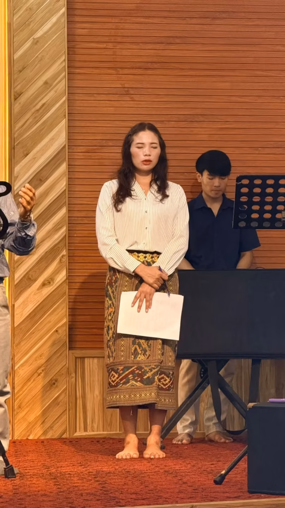 A woman in a striped blouse and woven skirt standing with eyes closed and a paper in hand on a church stage, with a young man behind her in Laos.