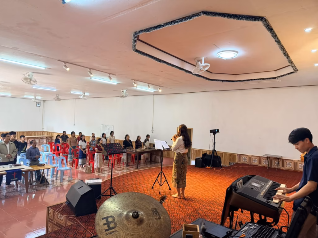 A woman singing into a microphone on a carpeted stage while a young man plays keyboard and a congregation stands in the hall beyond in Laos.