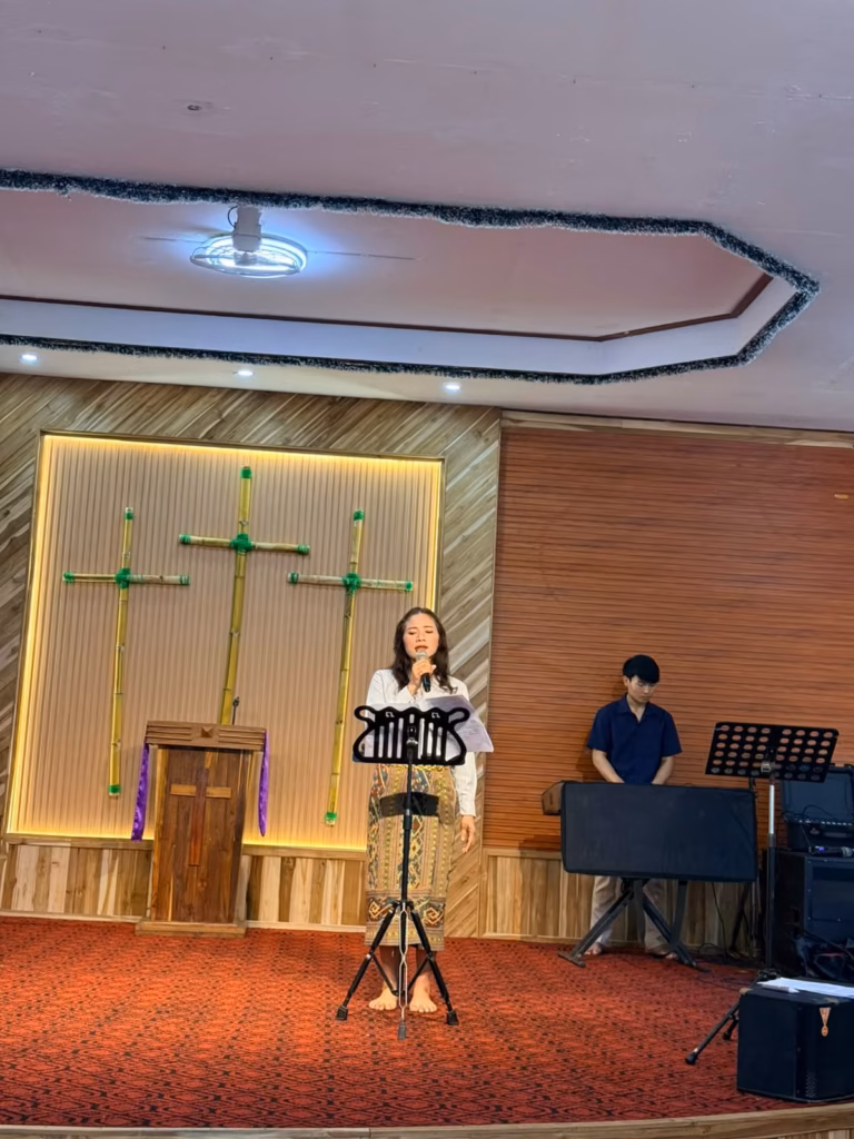 A woman singing into a microphone at a music stand on a church stage with three bamboo crosses and a wooden pulpit behind her in Laos.