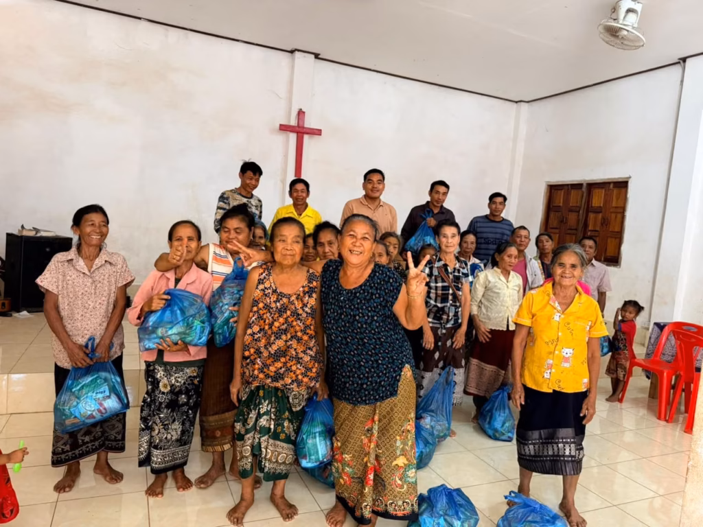 Women holding blue bags of supplies and smiling in front of a red cross on the wall of a church hall, with five men standing behind them in Laos. Pastor Khen is pictured.