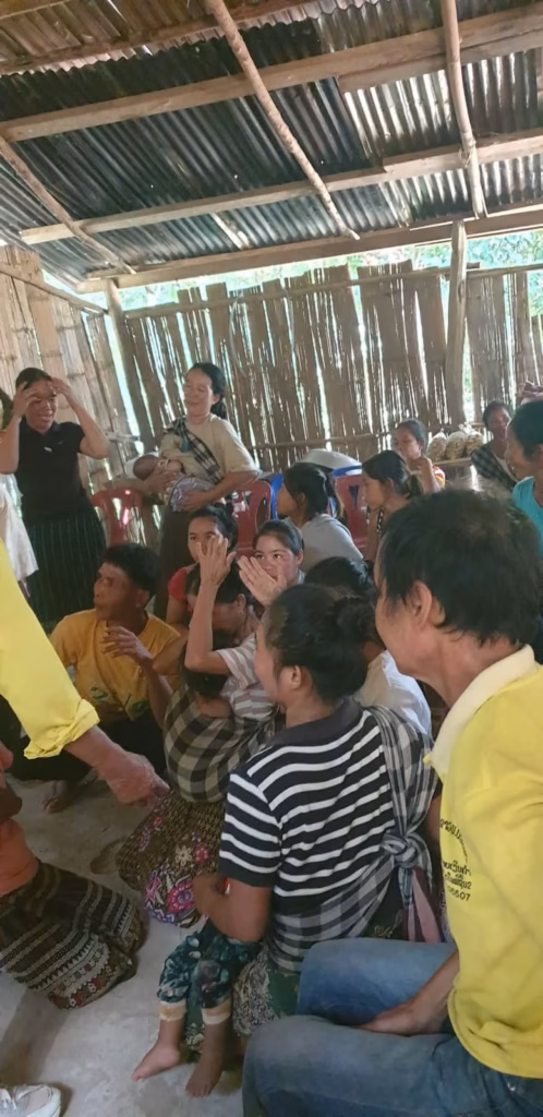 Women, men and children sitting on a concrete floor in an open bamboo-walled shelter, one woman standing and holding a baby in Laos.