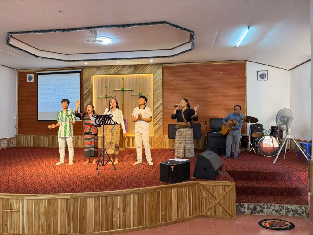 A pastor playing acoustic guitar beside four singers on a carpeted church stage with bamboo crosses and a lyrics screen in Laos.