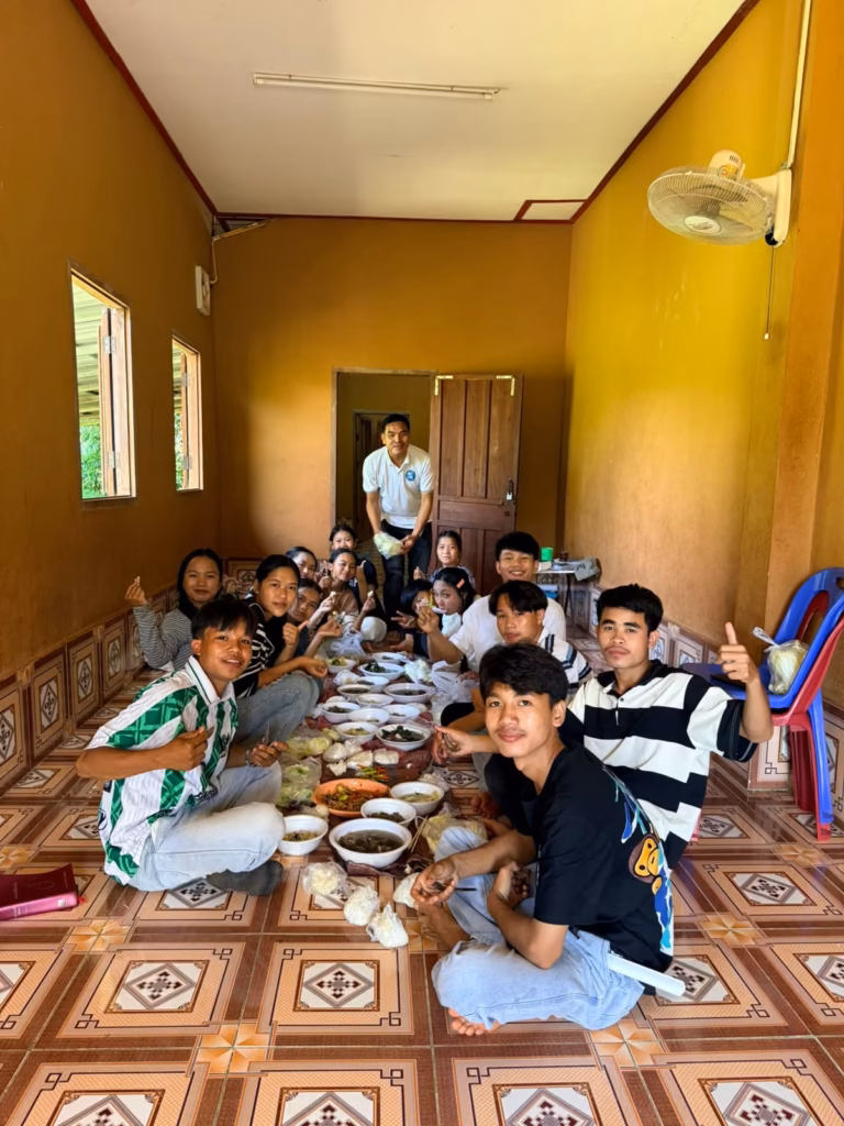 Pastor Khen standing at the back of a long room while young people sit on a tiled floor around bowls of food in Laos.